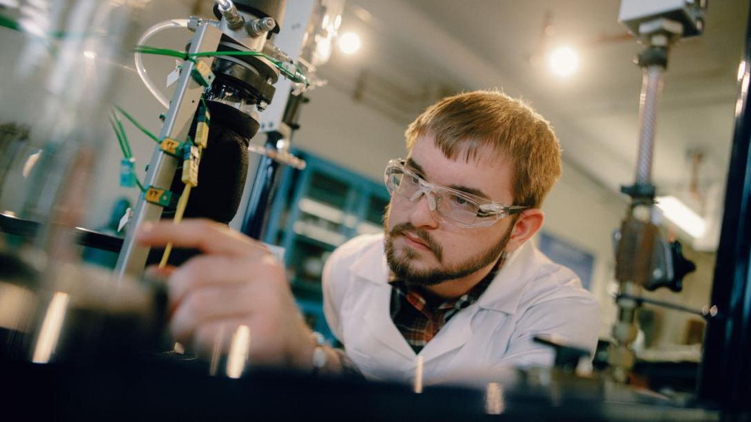 A Kettering chemical engineering student wears safety glasses and a white lab coat and inspects lab equipment